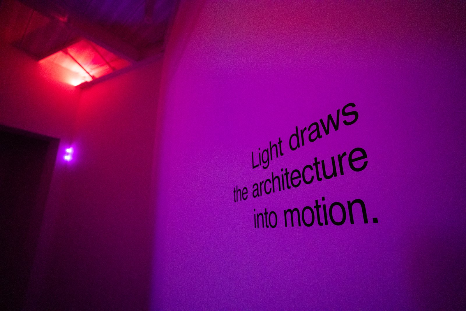 Magenta laser light illuminates the wall text, ‘Light draws the architecture into motion.’